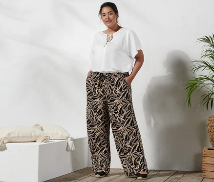 Une femme se tient debout, les mains dans les poches d'un pantalon palazzo imprimé noir et beige, portant un chemisier avec lien à nouer et des espadrilles à talon compensé.