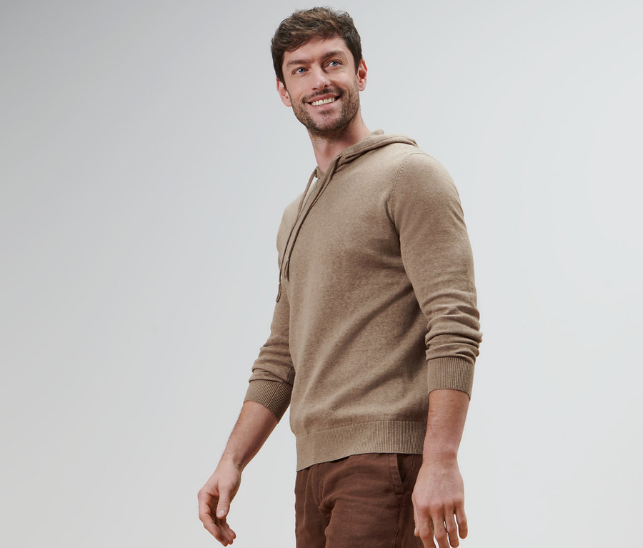 Un homme souriant portant un sweatshirt à capuche en maille et un pantalon marron.