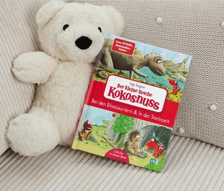 Un ours en peluche est posé à côté du livre «	Der kleine Drache Kokosnuss – Bei den Dinosauriern & In der Steinzeit	» (Le petit dragon Noix de coco – Chez les dinosaures et à l’âge de pierre, en allemand).