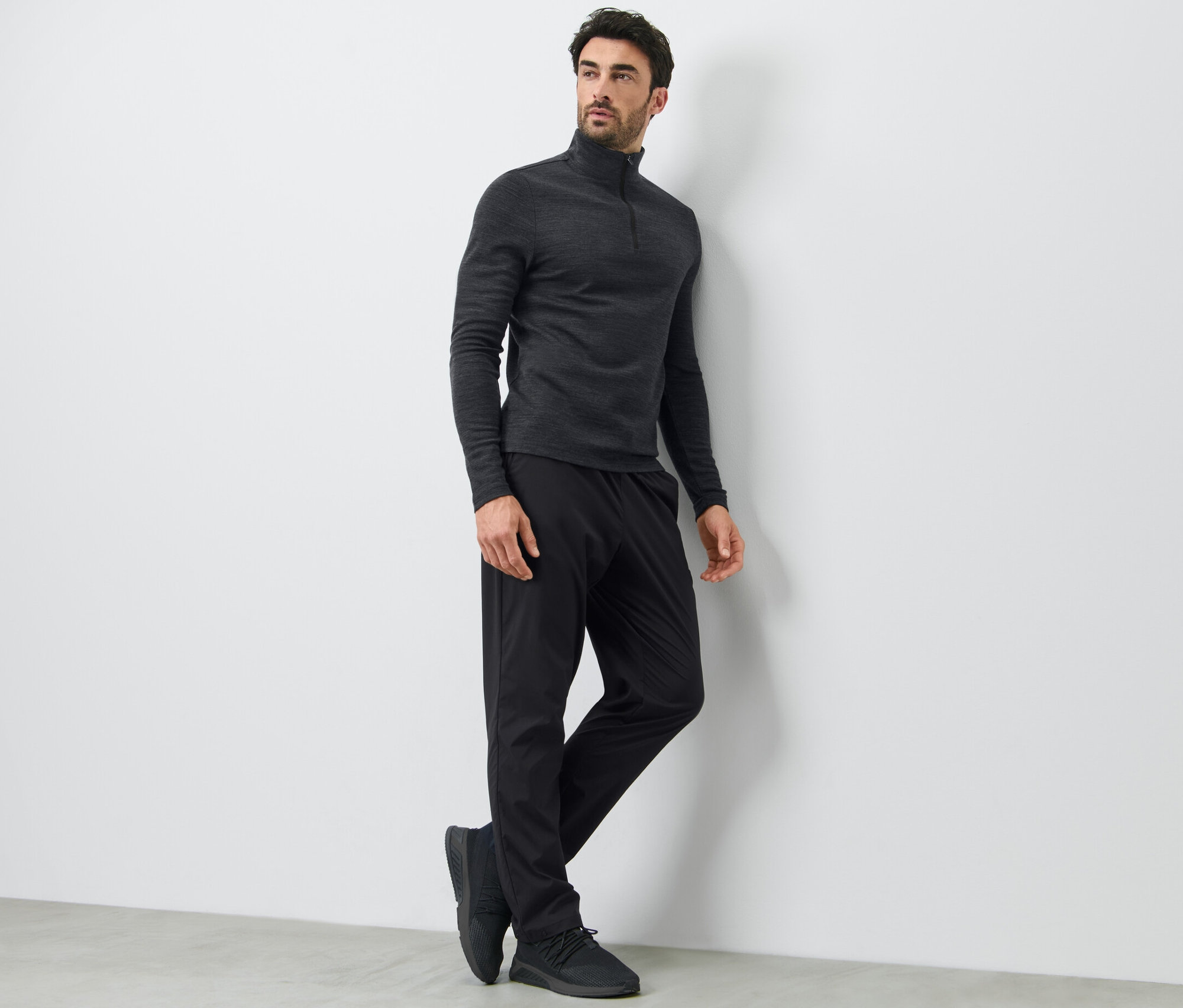 Un homme appuyé contre un mur porte un pantalon technique noir et un haut à manches longues.