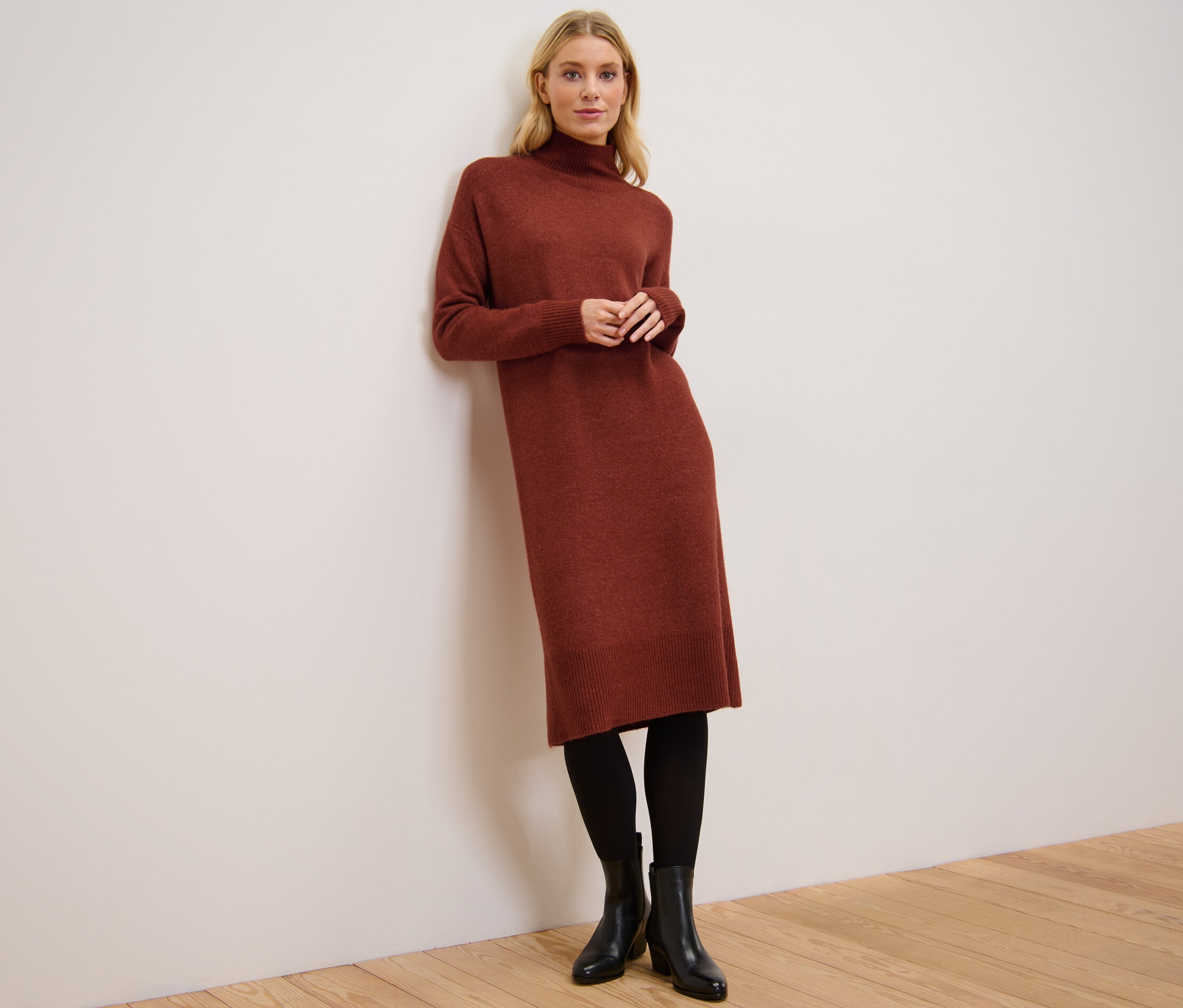 Femme appuyée contre un mur portant une robe en maille avec teneur en laine, des collants noirs et des bottines.