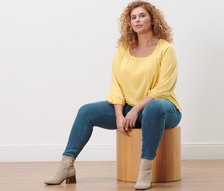 Une femme est assise sur un tabouret en portant une blouse jaune manches 3/4 et un tregging « Fit Nelly ».