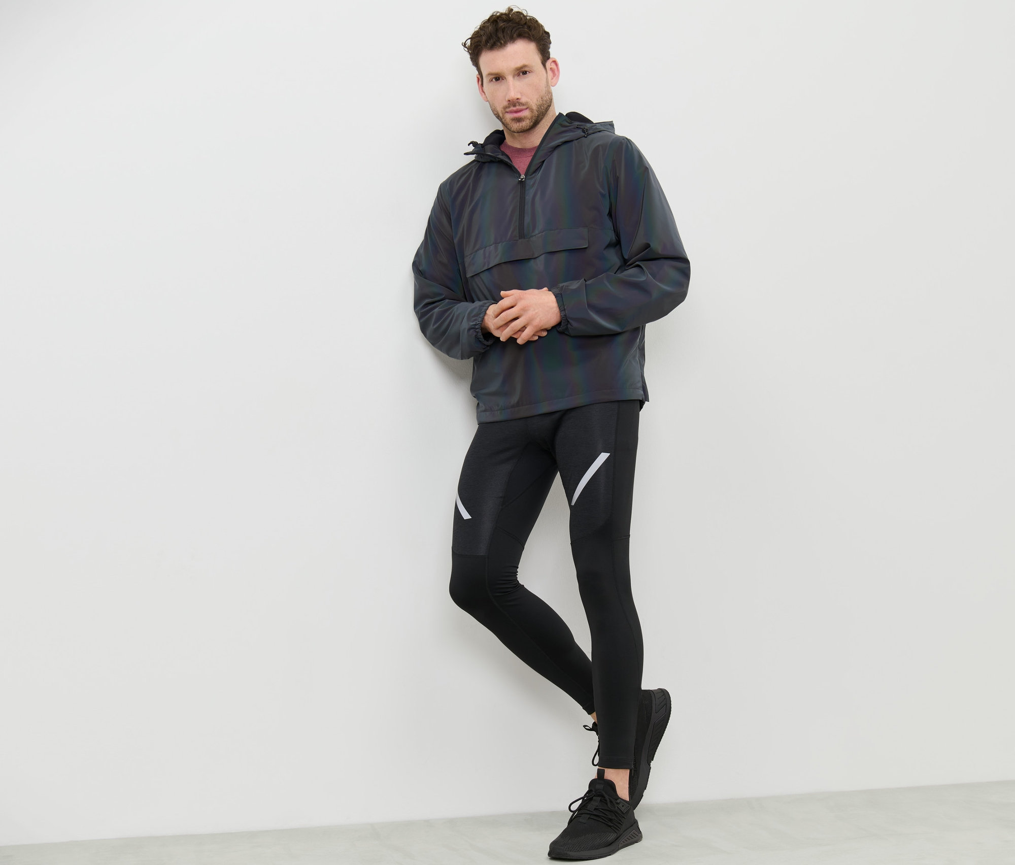 Un homme appuyé contre un mur portant un coupe-vent Rainbow Reflective, un legging noir et des baskets.