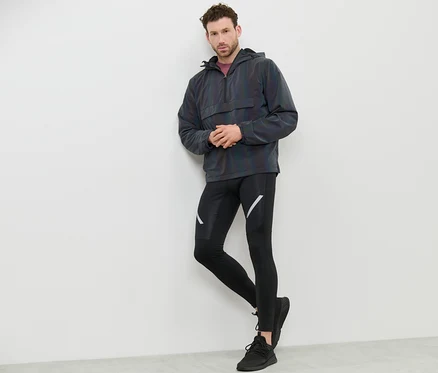 Un homme appuyé contre un mur portant un coupe-vent Rainbow Reflective, un legging noir et des baskets.