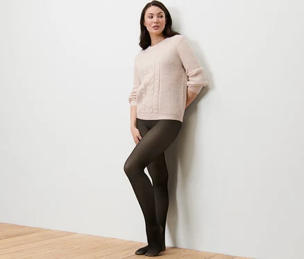 Femme appuyée contre un mur portant un pull et des collants fins doublés en aspect transparent.