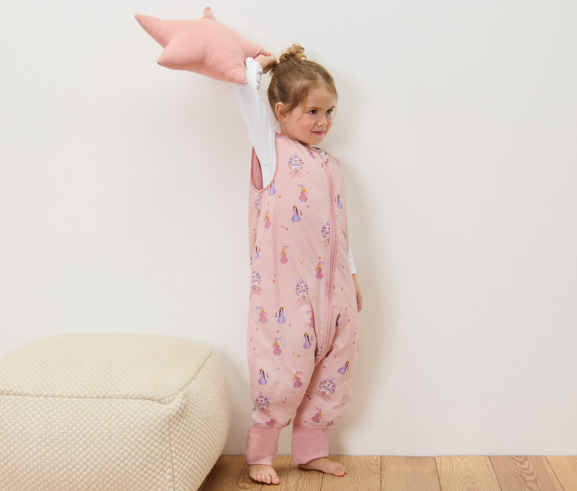 Une fille tient un coussin rose en forme d'étoile et porte une gigoteuse avec jambes rose de 2,5 TOG, env. 110 cm.
