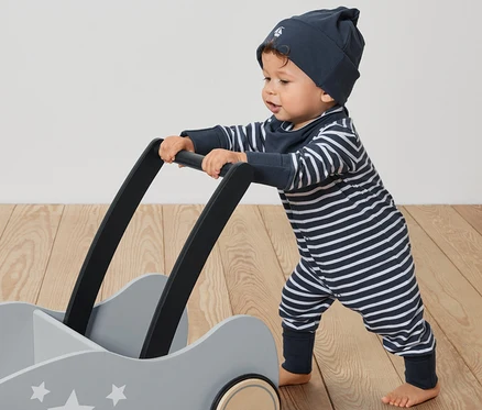 Un bébé pousse un chariot gris avec des étoiles. Il porte un ensemble pour bébé, 3 pièces : une combinaison rayée, un bonnet et un bavoir.