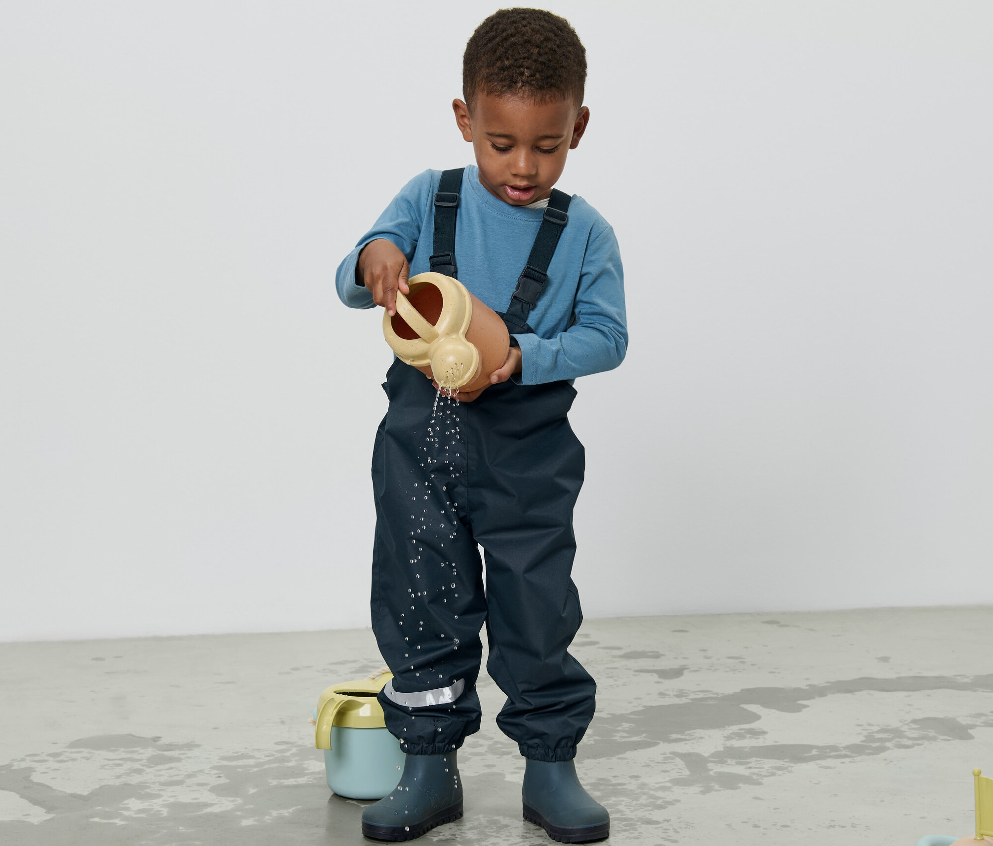 Un garçon en pantalon de pluie pour enfant verse de l'eau d'un arrosoir sur le sol.