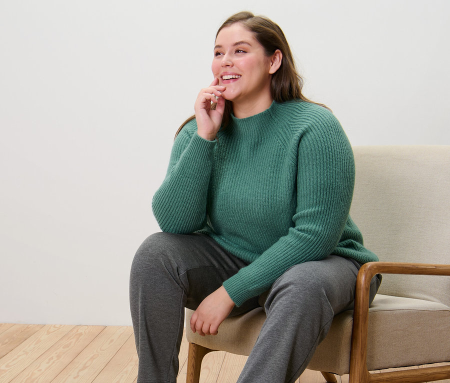 Une femme est assise sur une chaise et porte un pull à col droit en grosse maille vert et un pantalon de jogging.