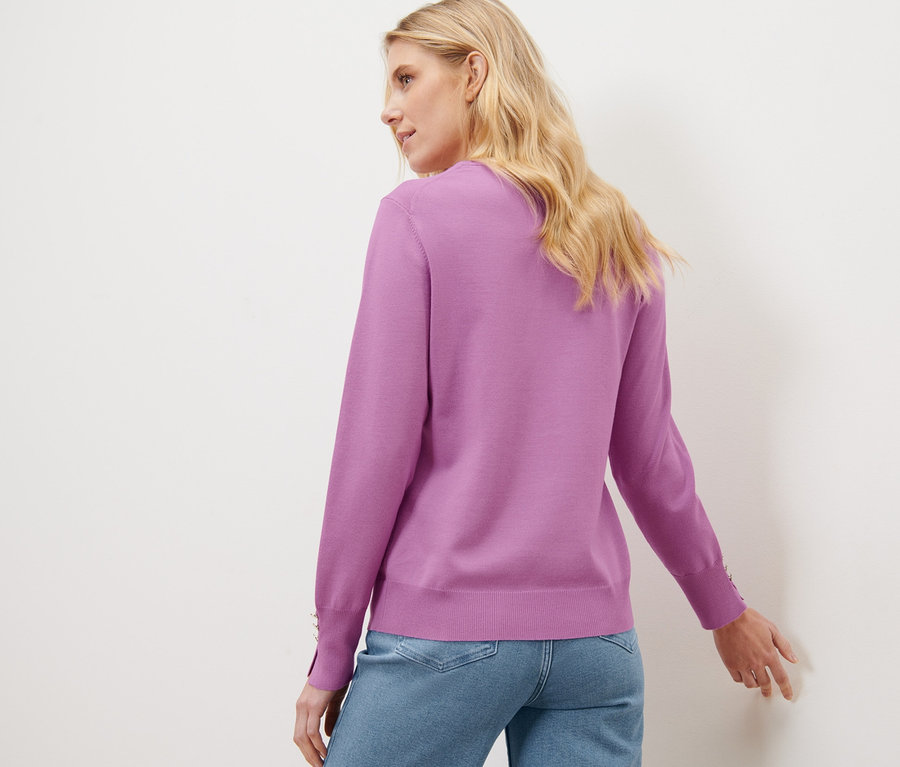 Une femme blonde pose dos à la caméra, portant un pull en maille fine violet et un jean.