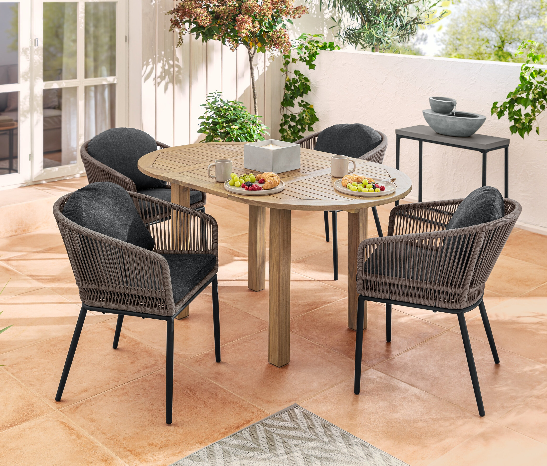 Quatre fauteuils de salon avec dossier arrondi et tressage textile autour d'une table ronde avec nourriture et boisson sur une terrasse. À l'arrière-plan, une fontaine de jardin solaire et un set de table console.