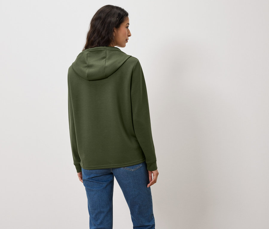 Une femme, dos à la caméra, porte un sweat à capuche vert et un jean.