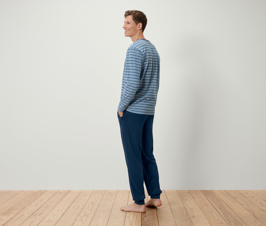 Un homme se tient de côté, portant un t-shirt à manches longues bleu rayé et un pantalon bleu foncé.