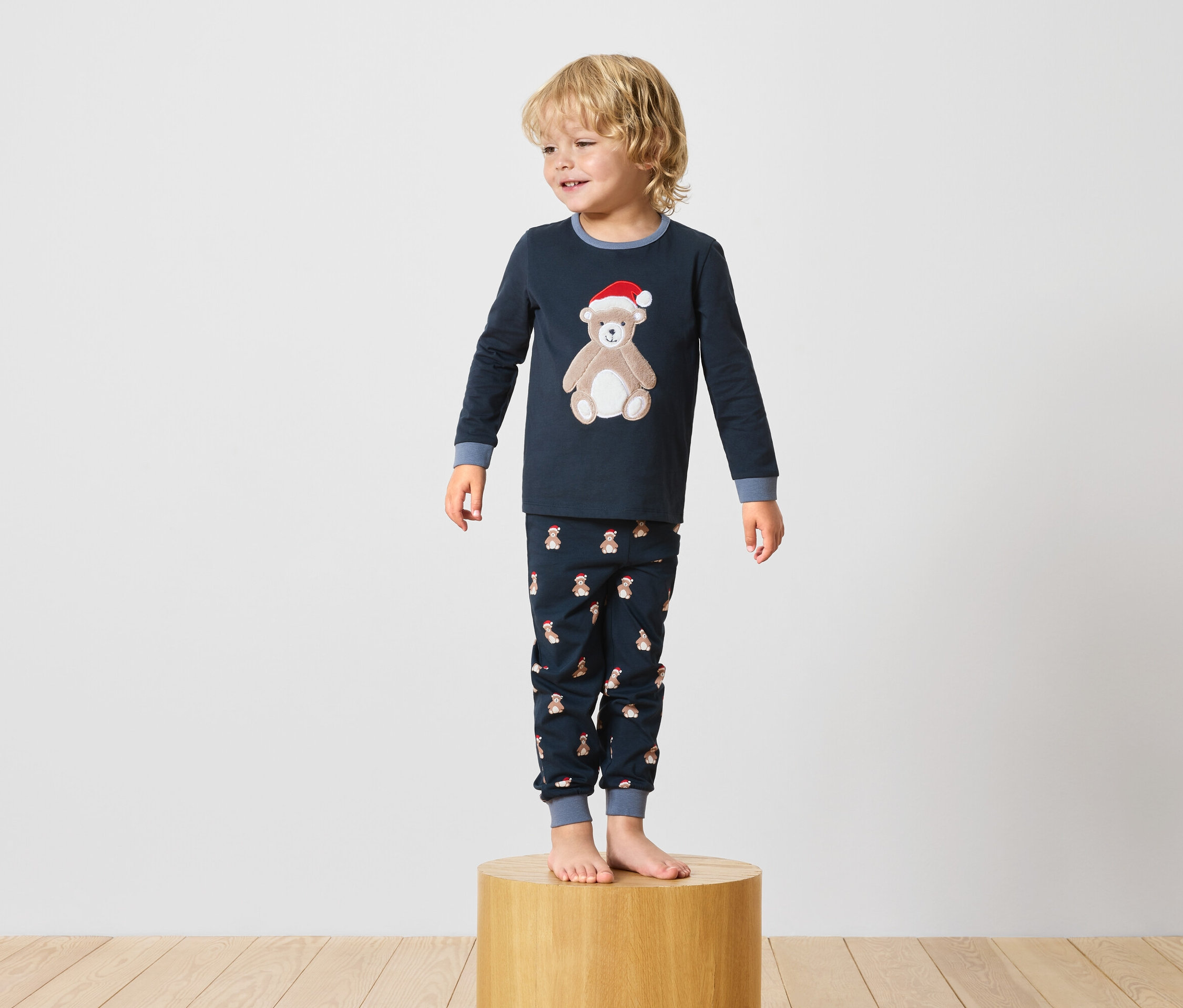 Un garçon se tient sur un socle en bois et porte un pyjama bleu marine avec un motif d'ours en peluche.