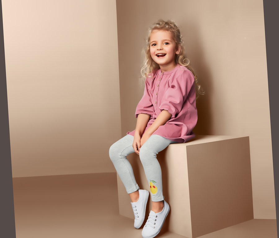 Une fillette souriante est assise sur un cube et porte 2 leggings avec une image de citron et des baskets blanches.