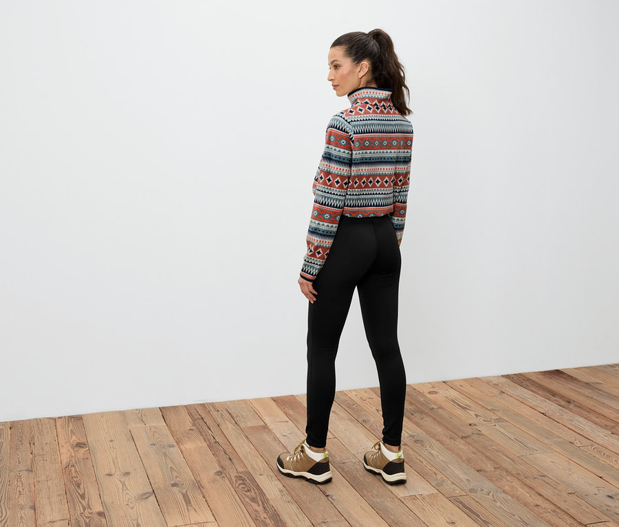 Une femme brune aux cheveux attachés en queue de cheval, vue de dos, porte un haut à manches longues à motifs, un legging noir et des chaussures marron.