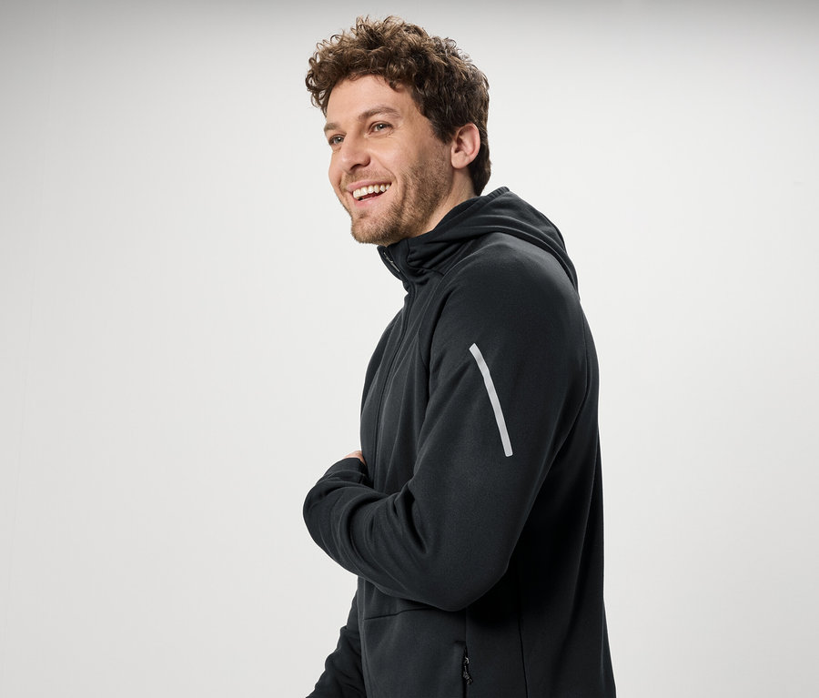 Un homme souriant aux cheveux bouclés porte un sweat à capuche noir.