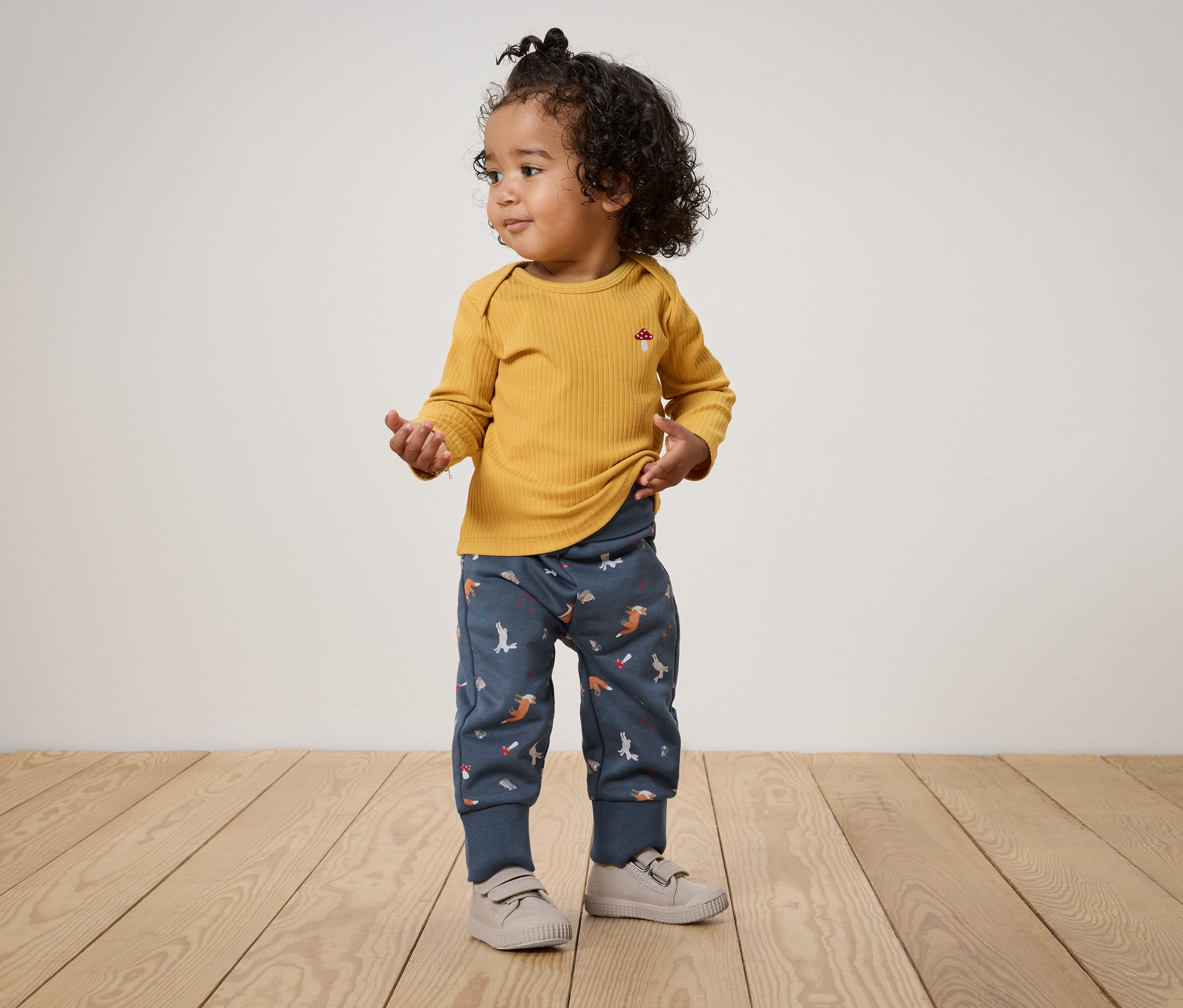 Un enfant aux cheveux bouclés se tient sur un plancher en bois, portant un t-shirt à manches longues jaune moutarde et un pantalon bleu avec des imprimés d'animaux.