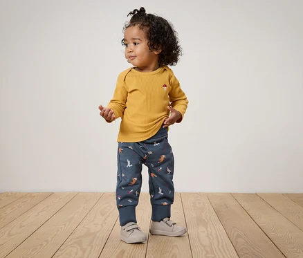 Un enfant aux cheveux bouclés se tient sur un plancher en bois, portant un t-shirt à manches longues jaune moutarde et un pantalon bleu avec des imprimés d'animaux.
