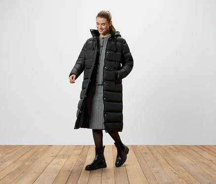 Une femme portant une longue doudoune noire, une robe tricotée grise et des bottes noires se tient sur un plancher en bois.