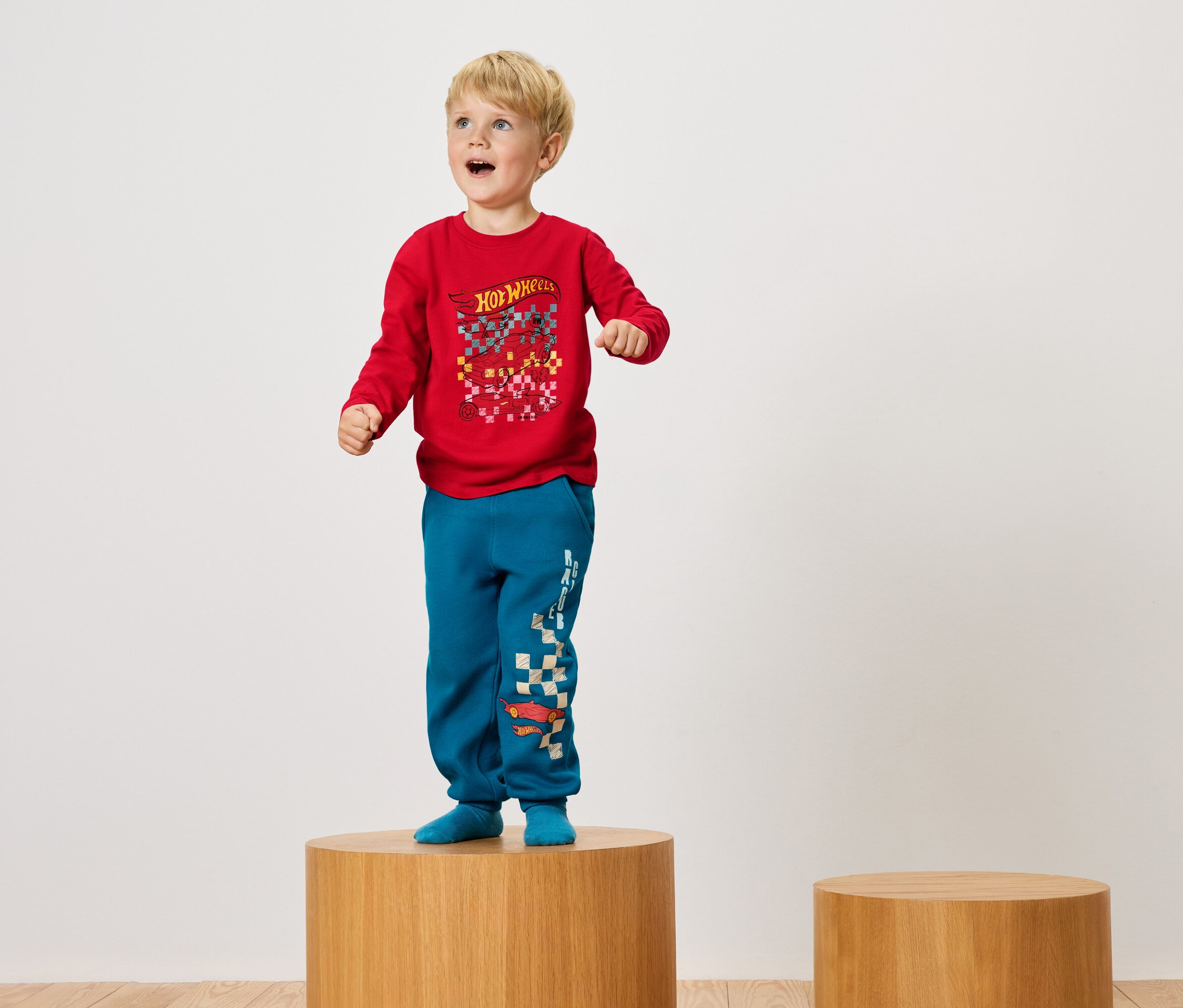 Un garçon debout sur un socle en bois, portant un t-shirt rouge et un pantalon bleu.
