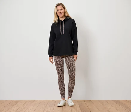 Une femme debout sur un plancher en bois porte un sweat à capuche noir et des leggings à motif animal.