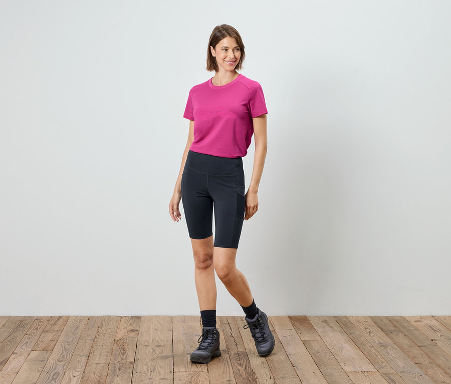 Une femme se tient sur un plancher en bois et porte un t-shirt rose, un short de cycliste noir, des chaussettes et des chaussures de randonnée.