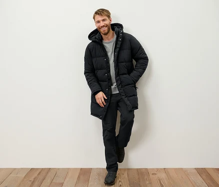 Un homme souriant avec une barbe est appuyé contre un mur et porte une doudoune noire à capuche, un pantalon noir et des chaussures.