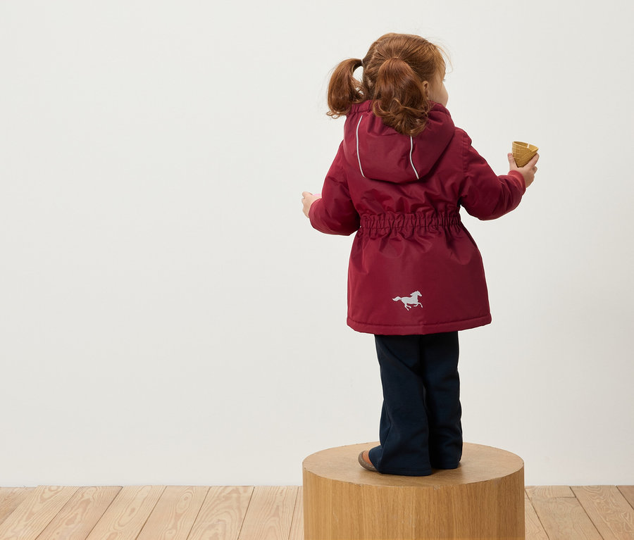 Une fille rousse avec des couettes se tient dos à l'appareil photo sur un socle en bois, porte une veste bordeaux à capuche avec une image de cheval et tient un cornet de glace à la main.