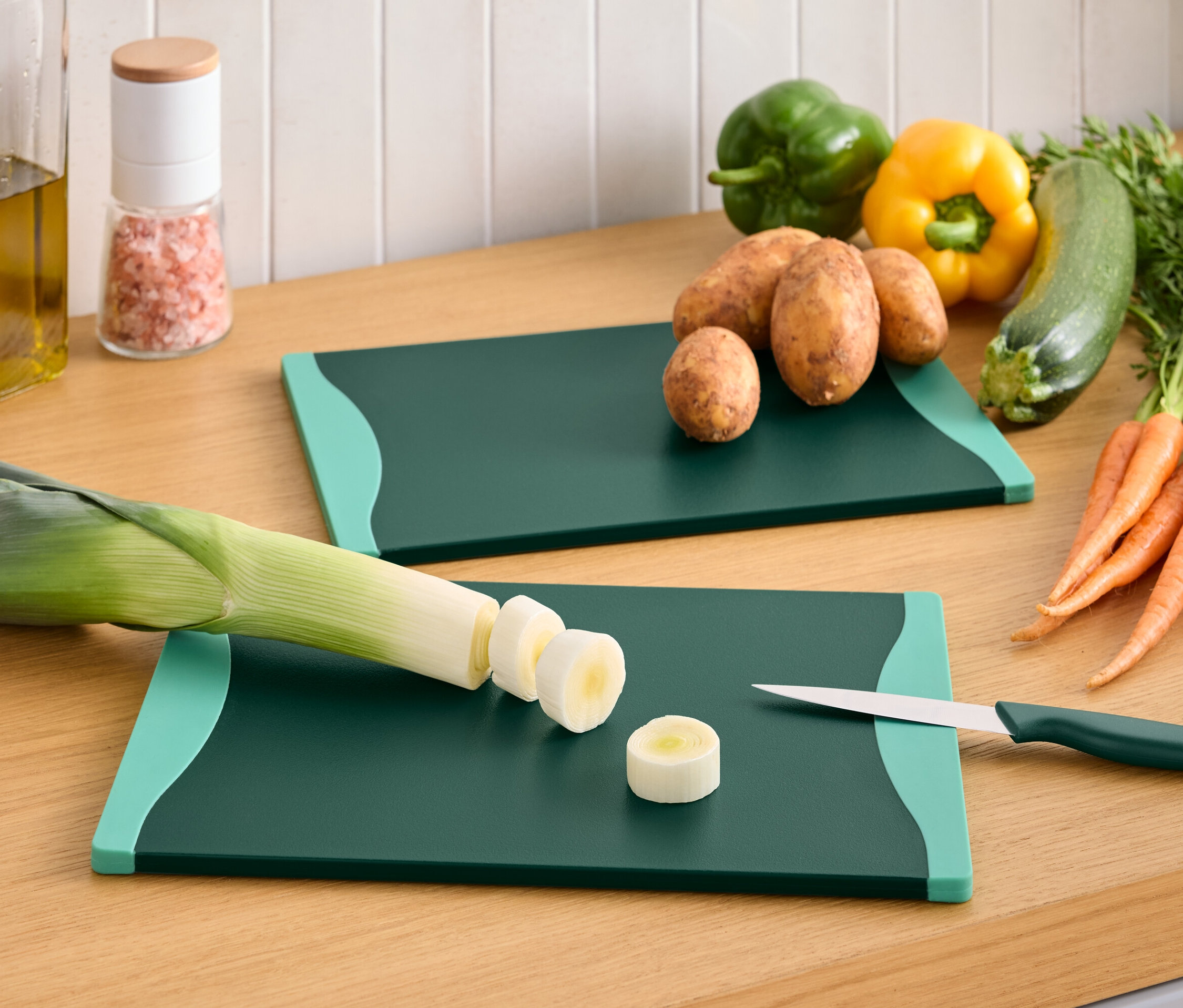 Sur une surface en bois, il y a deux planches à découper vertes avec différents types de légumes, notamment du poireau, des poivrons, des pommes de terre, des courgettes et des carottes.