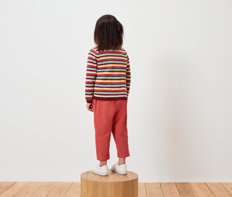 Un enfant dos à la caméra se tient sur un socle en bois, vêtu d'un pull rayé et d'un pantalon orange.