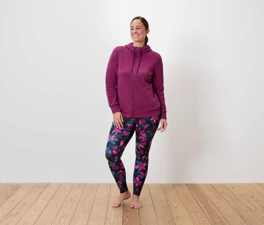 Femme portant un sweat à capuche bordeaux zippé et un legging à fleurs, debout sur un plancher en bois.