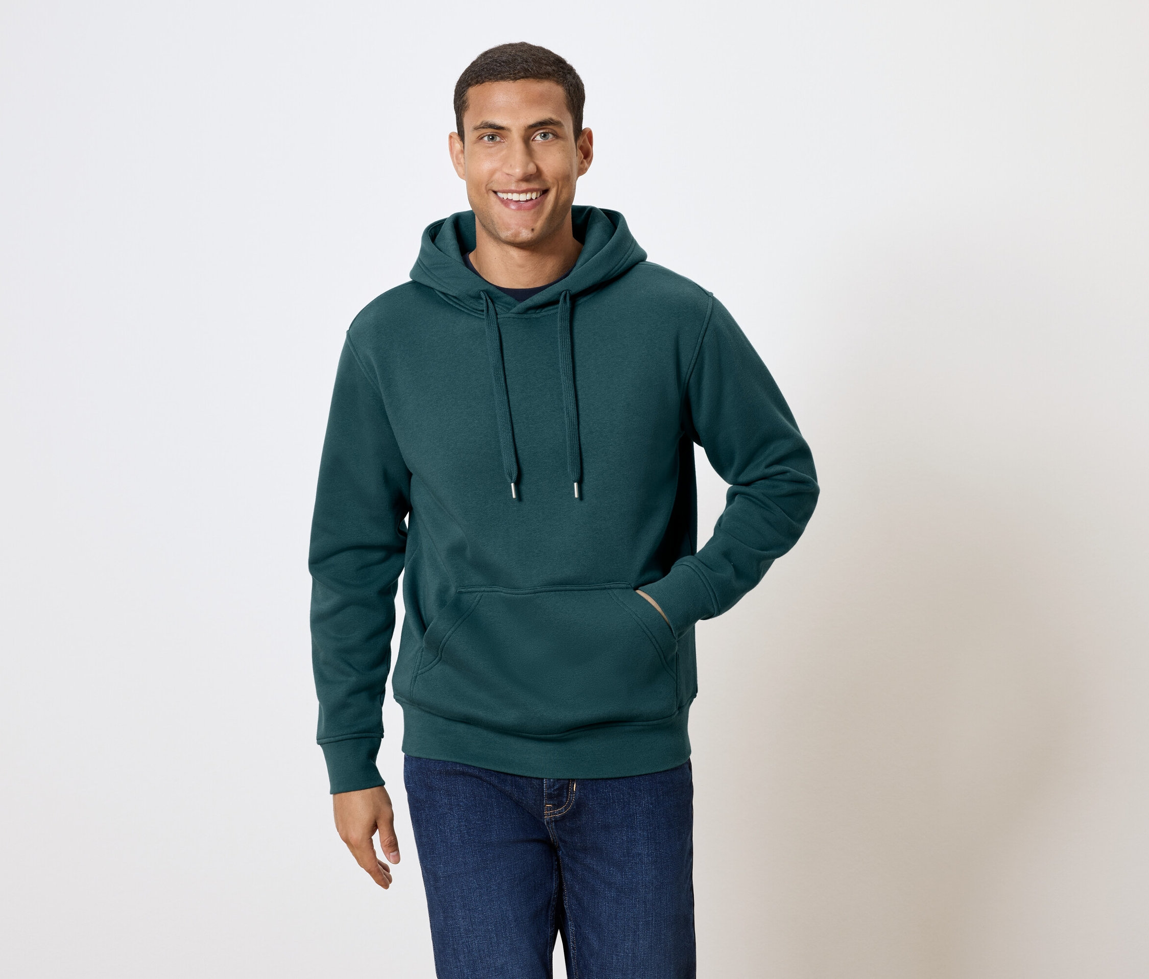 Un homme aux cheveux courts sourit et pose en sweat à capuche vert foncé et en jean bleu.