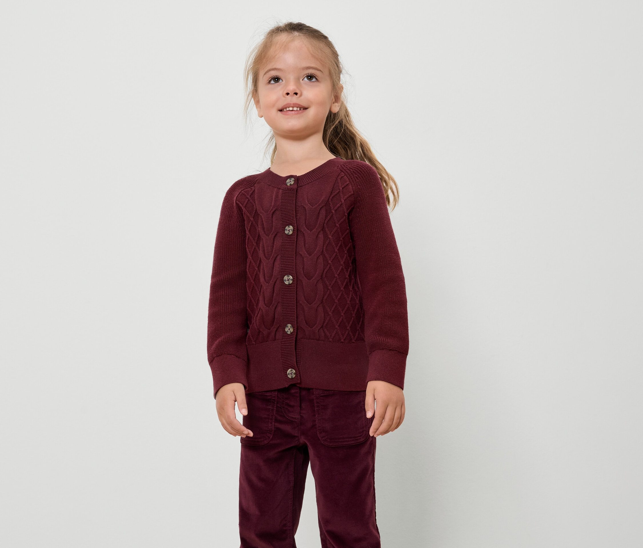 Une fille portant un cardigan et un pantalon bordeaux se tient devant un fond blanc.