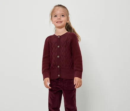 Une fille portant un cardigan et un pantalon bordeaux se tient devant un fond blanc.