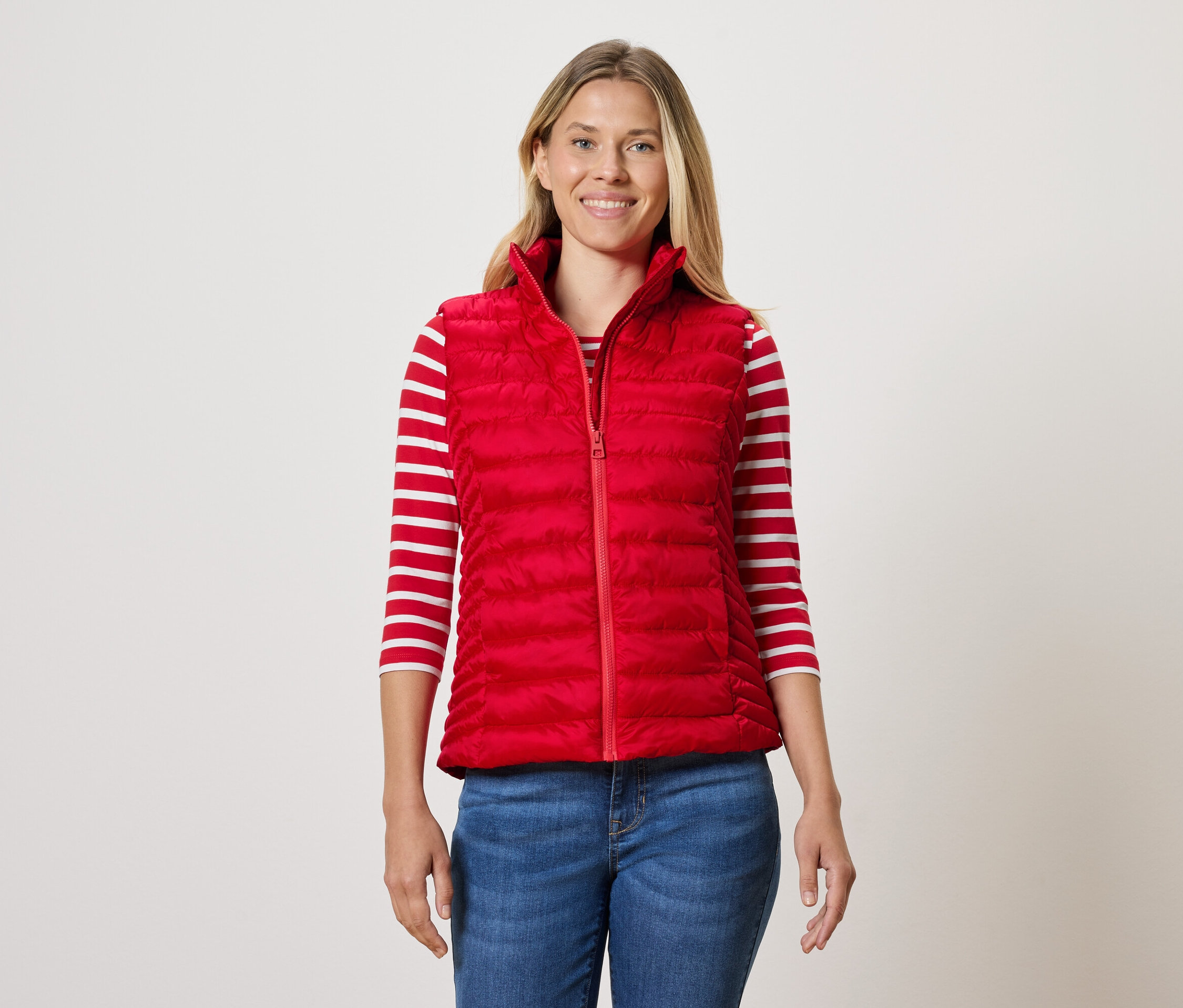 Une femme porte une veste matelassée rouge, un T-shirt rayé et un jean.