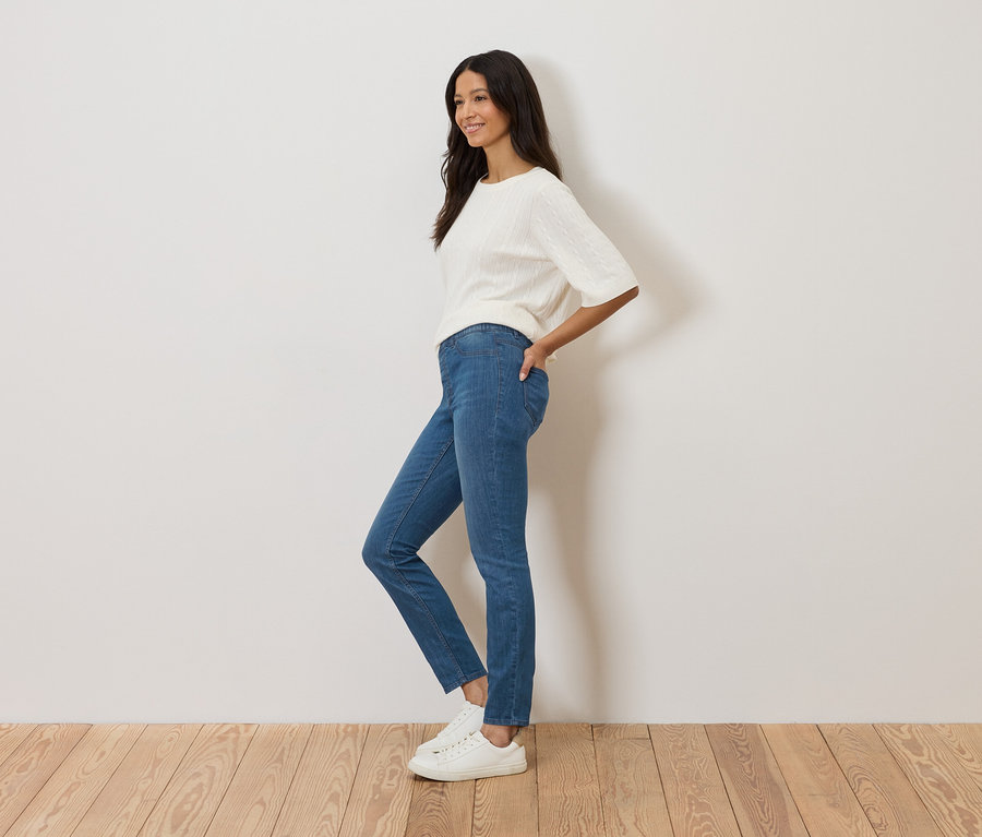 Une femme se tient de profil devant un mur blanc, porte un jean bleu, un pull blanc et des baskets blanches.