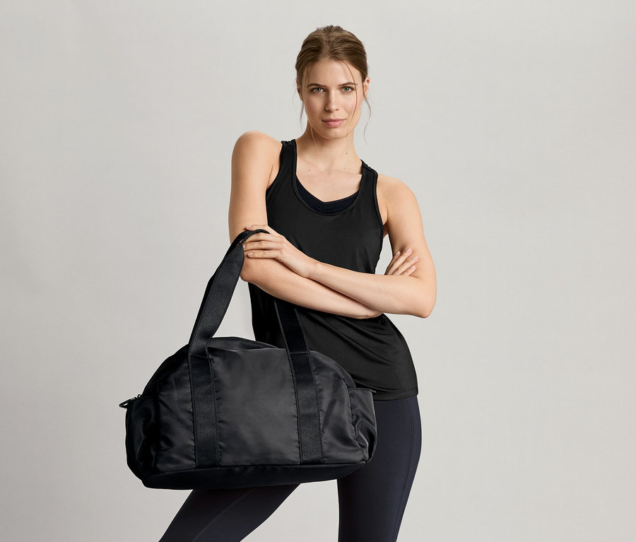 Femme les bras croisés tenant un sac de sport noir, portant un bustier de sport sans coutures noir et 2 hauts fonctionnels noirs et blancs.