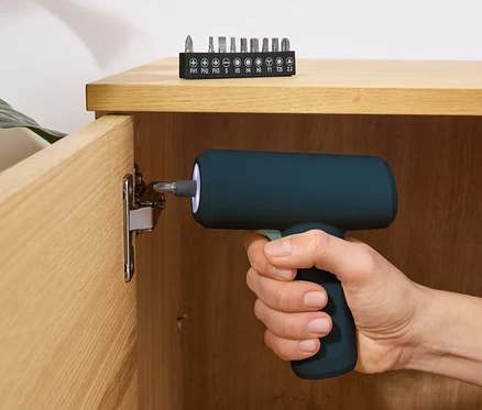 Une main tient un mini tournevis sans fil bleu 3,6 V et visse une vis dans une armoire en bois. Un jeu d'embouts repose sur le dessus de l'armoire.
