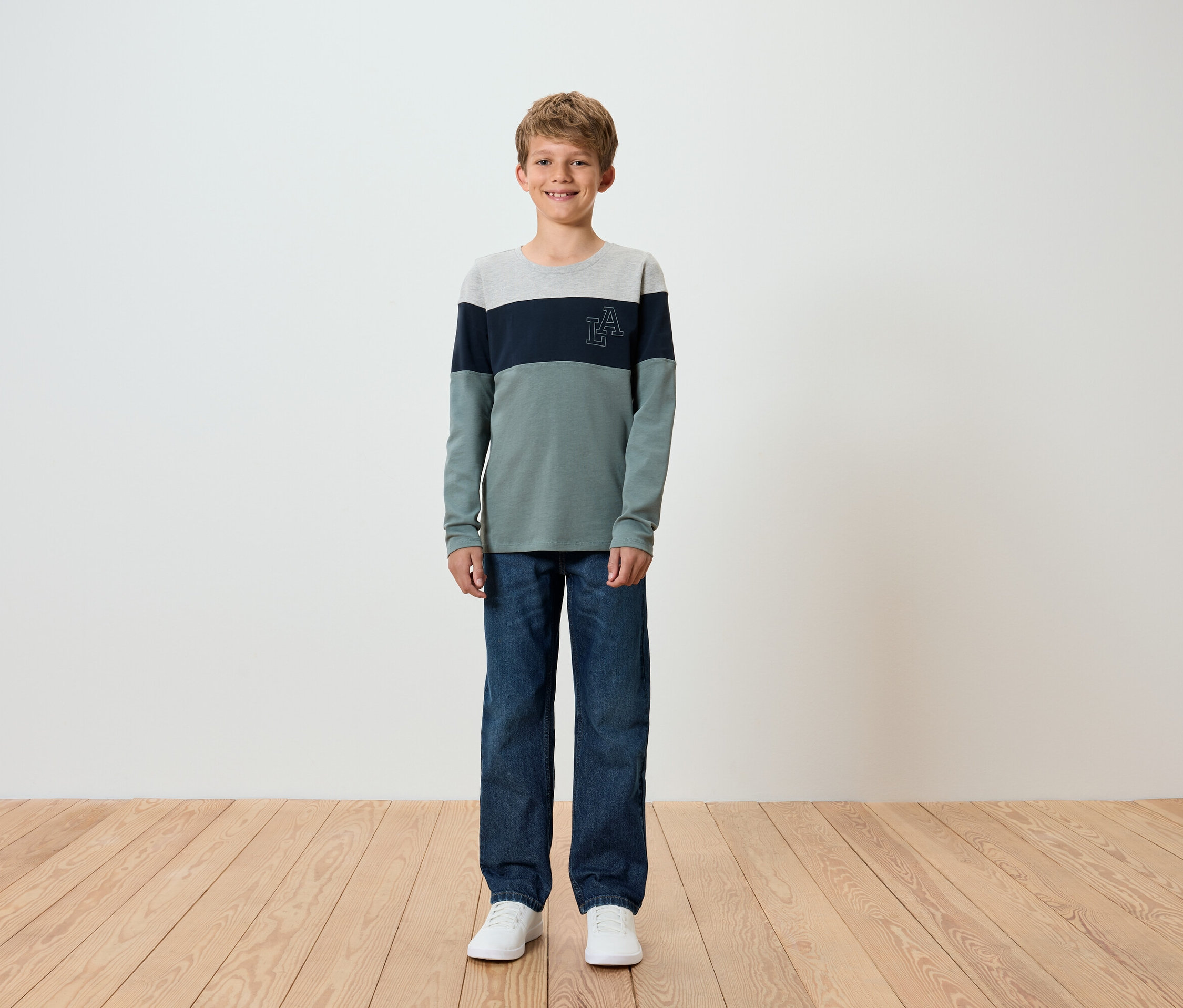 Un garçon debout sur un plancher en bois, portant un jean bleu et un t-shirt à manches longues.