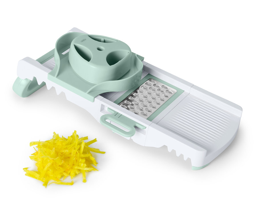 Une mandoline blanche avec des accents vert pastel à côté d'un tas de légumes jaunes râpés.
