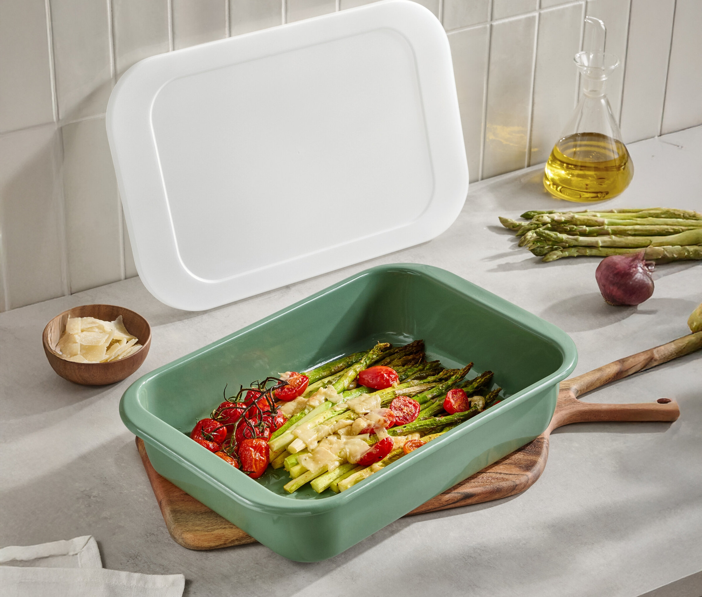Plat de cuisson vert avec asperges et tomates sur une table en bois. Un couvercle blanc, de l'huile d'olive, du parmesan et des oignons sont également visibles.