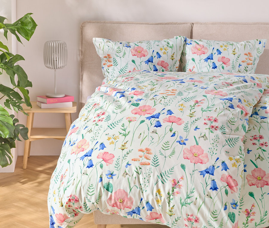 Lit avec des draps à motifs floraux, à côté une table avec une lampe et des livres, à gauche une plante.