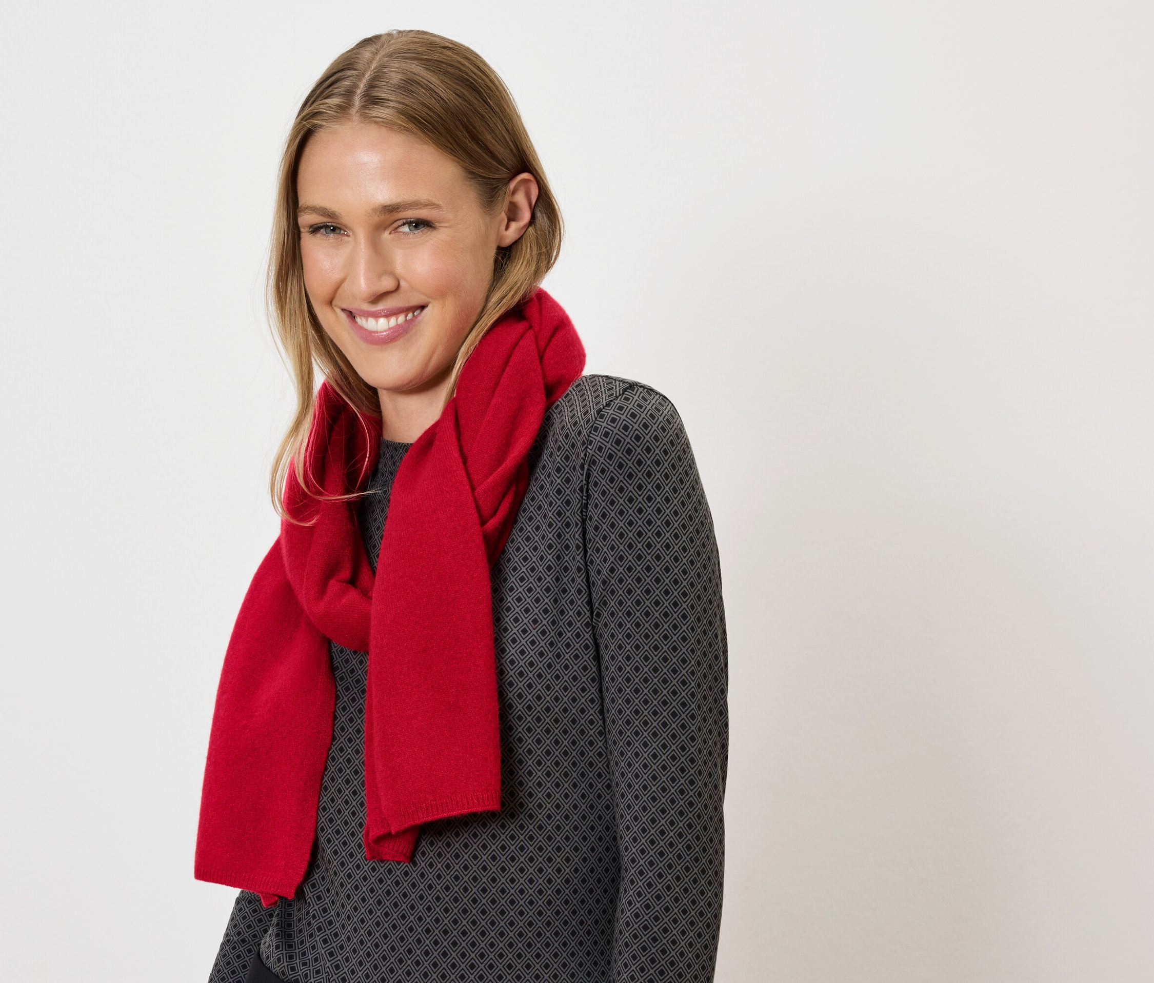 Femme souriante aux cheveux blonds et une écharpe rouge autour du cou sur un pull gris.