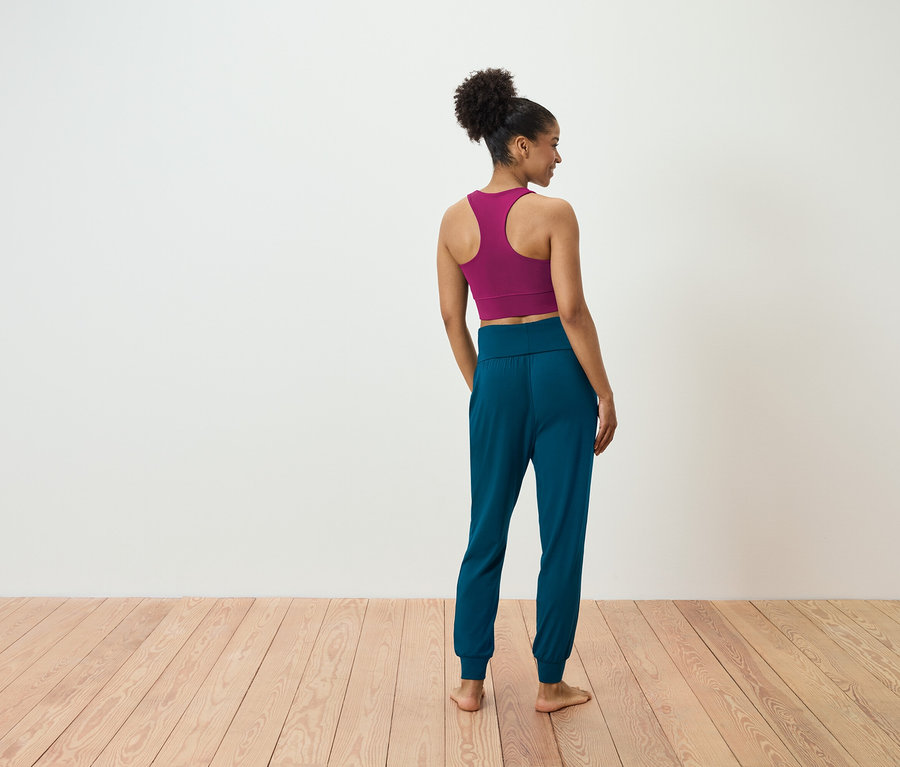 Une femme se tient dos à la caméra et porte un soutien-gorge de sport magenta et un pantalon de jogging bleu-vert.