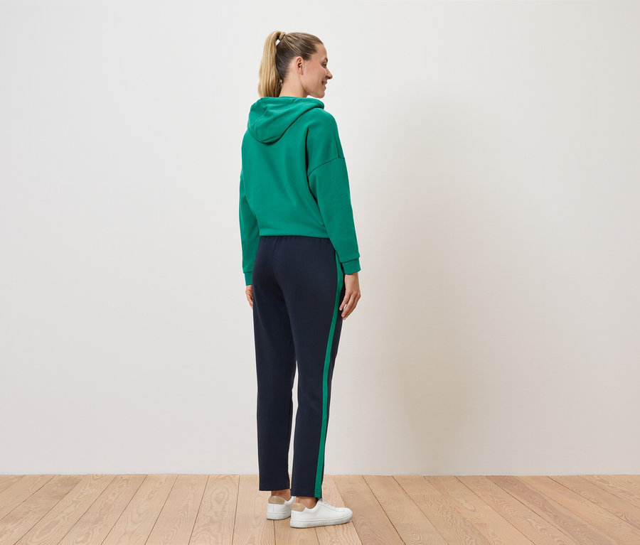 Une femme se tient dos à la caméra, portant un sweat à capuche vert et un pantalon bleu marine avec une bande.