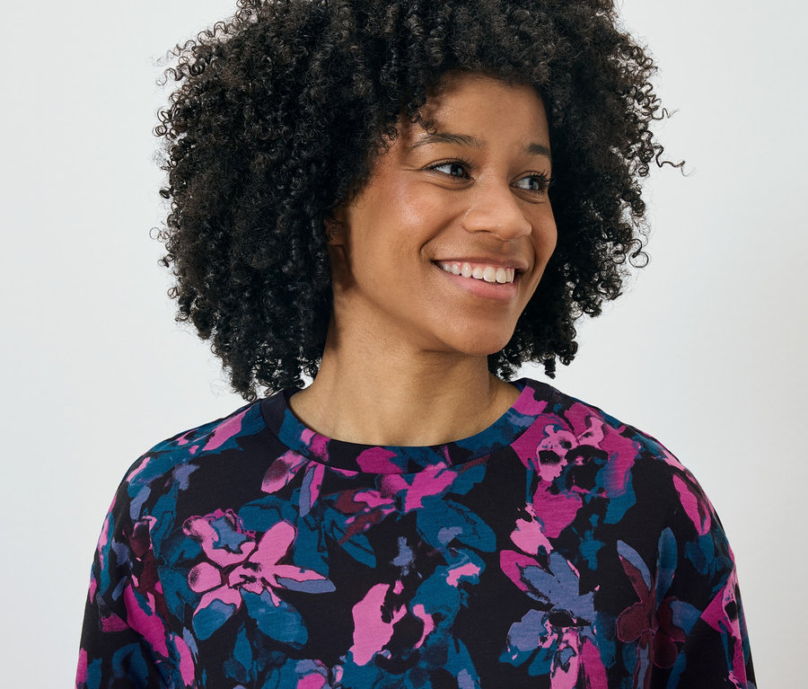 Portrait d'une femme souriante avec une coiffure afro portant un haut noir avec un motif floral rose et bleu.