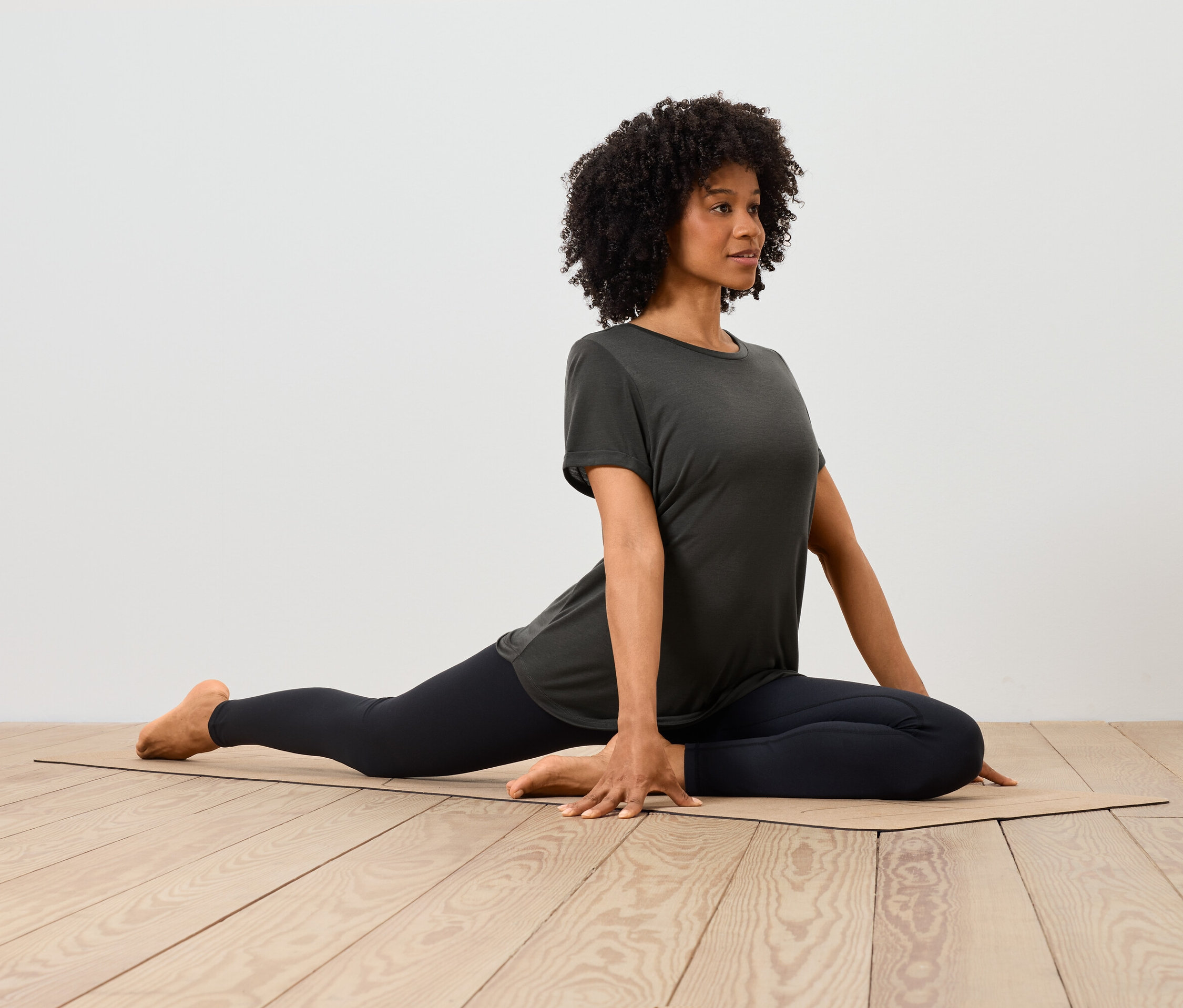Une femme afro-américaine en t-shirt et leggings s'étire sur un tapis sur un parquet.