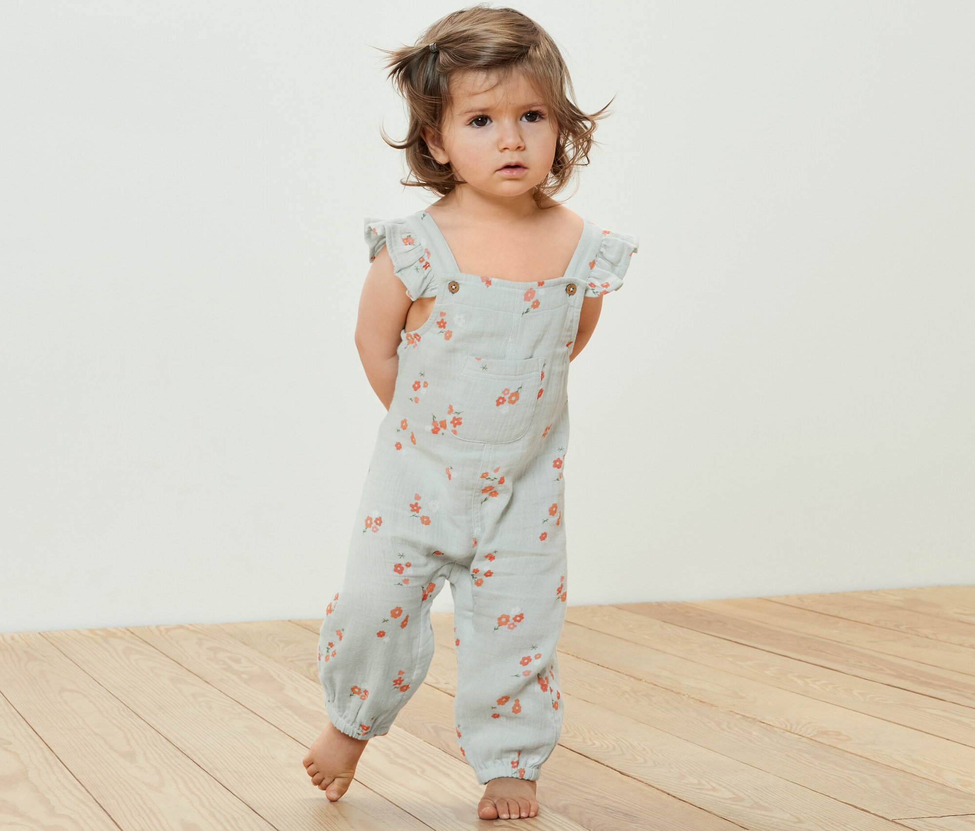 Un bébé se tient sur un plancher en bois et porte une barboteuse en mousseline pour enfant avec un motif floral.