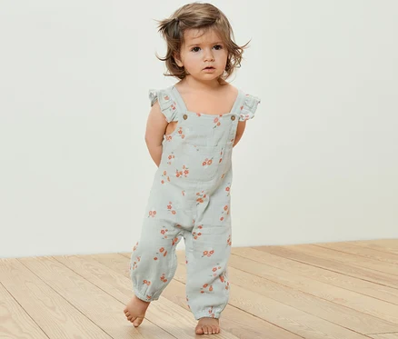 Un bébé se tient sur un plancher en bois et porte une barboteuse en mousseline pour enfant avec un motif floral.